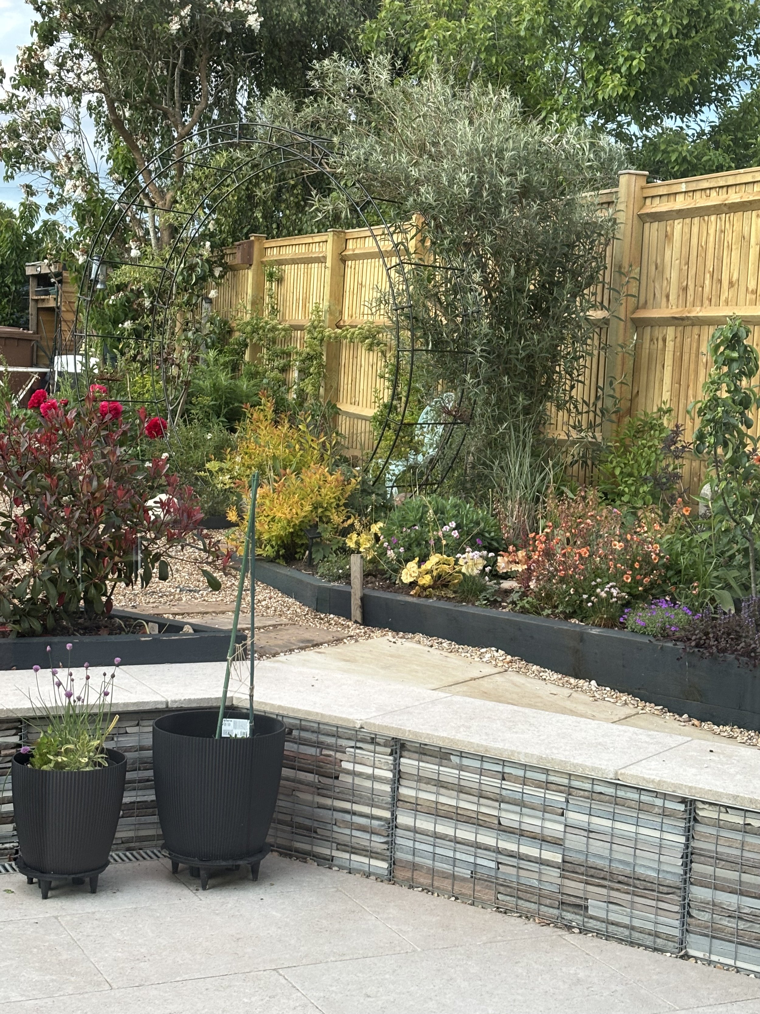 Garden with potted plants, flowers, a metal arch, a wooden fence, and trees.