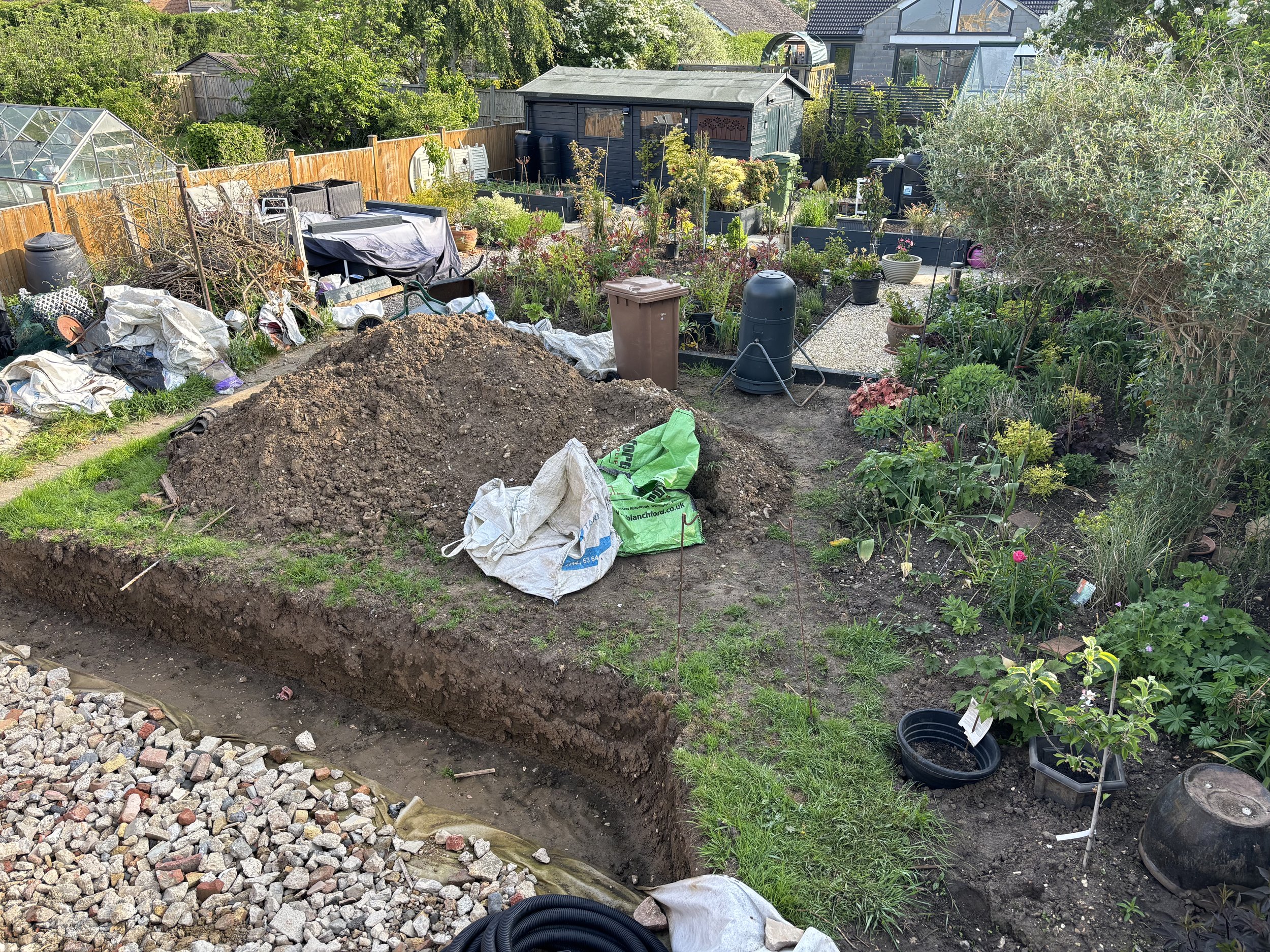 A backyard garden in the process of being landscaped, with a large pile of dirt, gardening tools, plants, and a pathway, enclosed by a wooden fence and houses in the background.