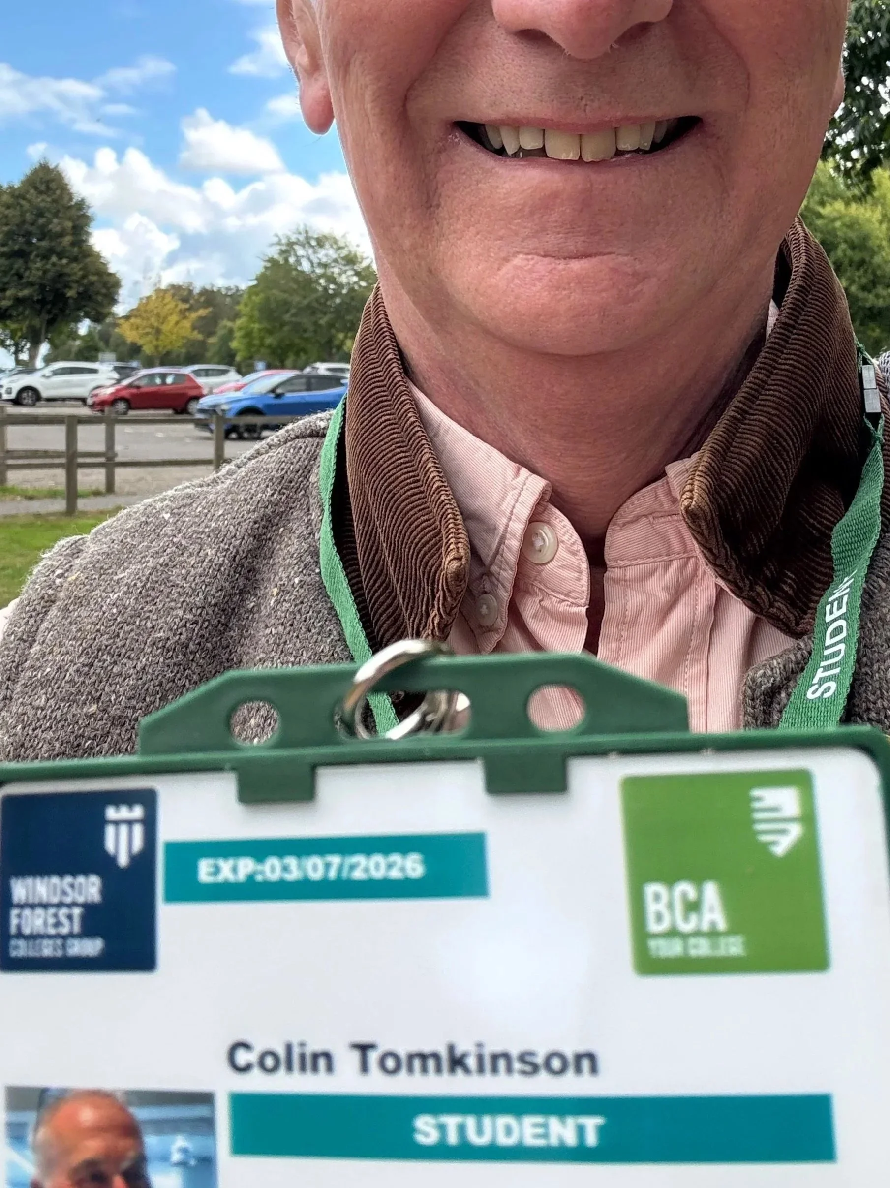 Close-up of a smiling man outdoors, with a parking lot and trees in the background. He is wearing a pink shirt and a gray jacket, holding a green clip with an identification badge visible, which displays the name "Colin Tomkinson" and indicates he is a student.