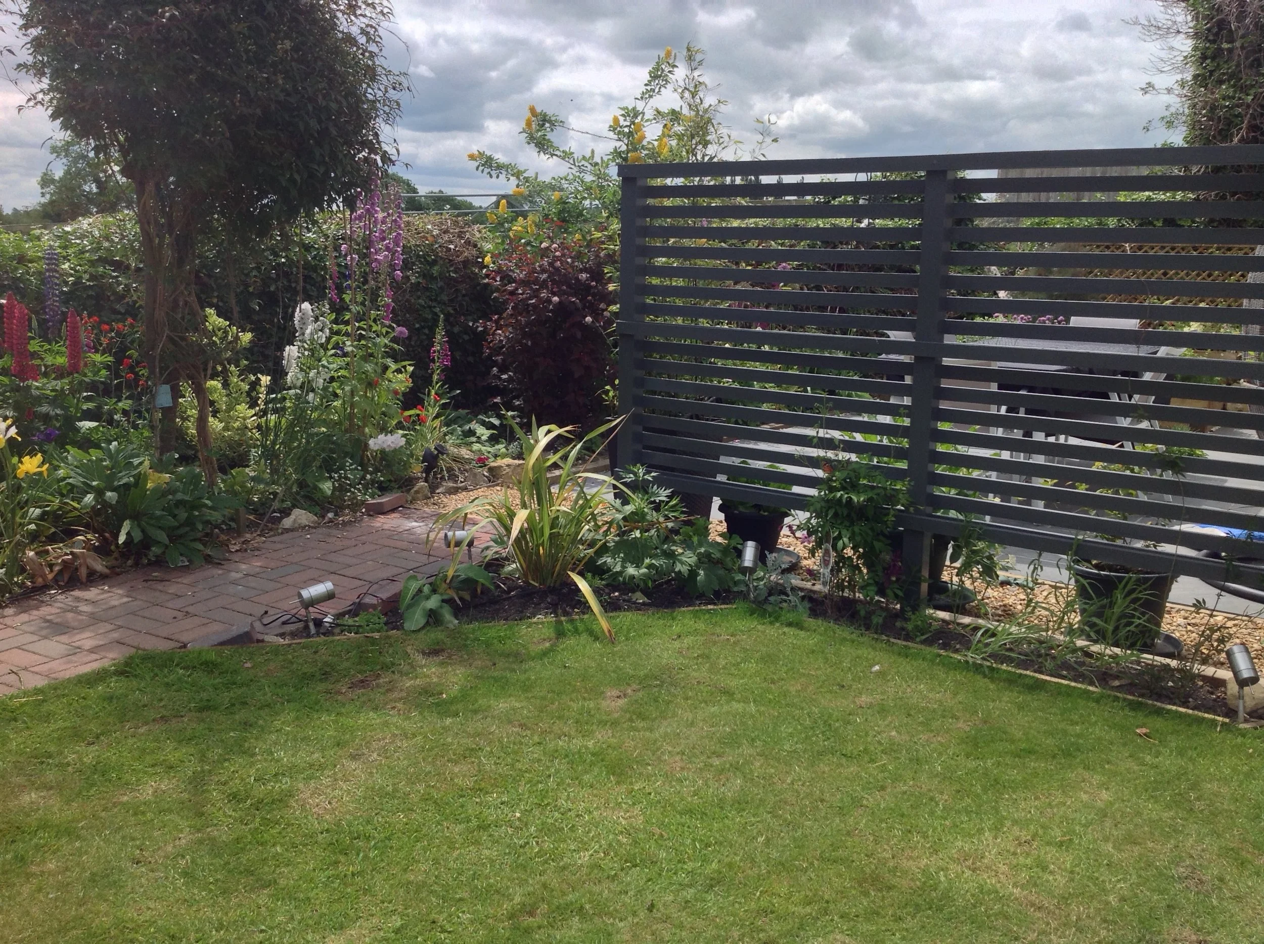 A garden with colorful flowers, a brick pathway, and green lawn in front of a black modern fence with horizontal slats under a partly cloudy sky.