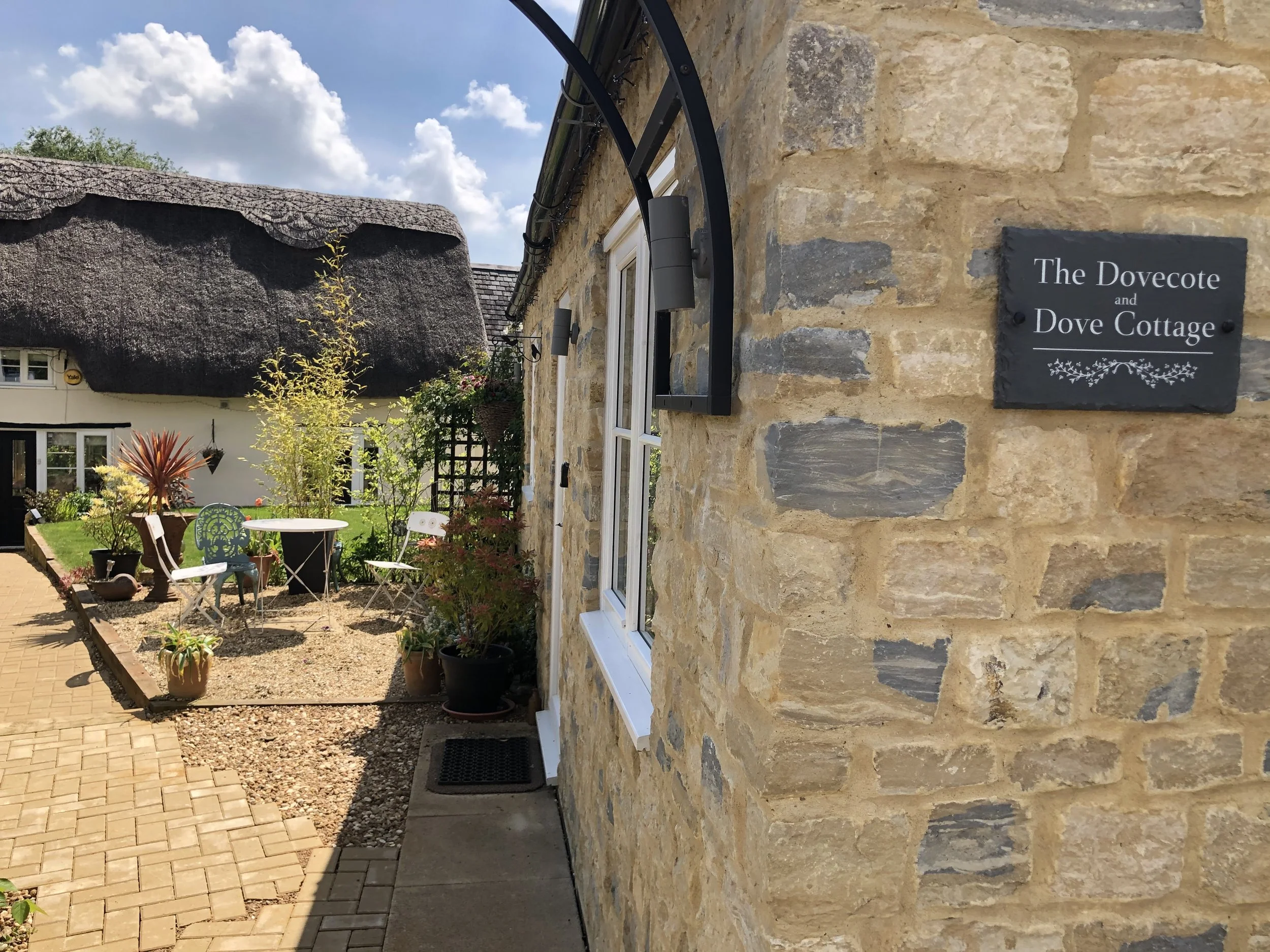 A stone building with a sign that reads 'The Dovecote and Dove Cottage' hangs on the wall. There is a garden area with outdoor furniture and potted plants in front of the building, under a partly cloudy sky.
