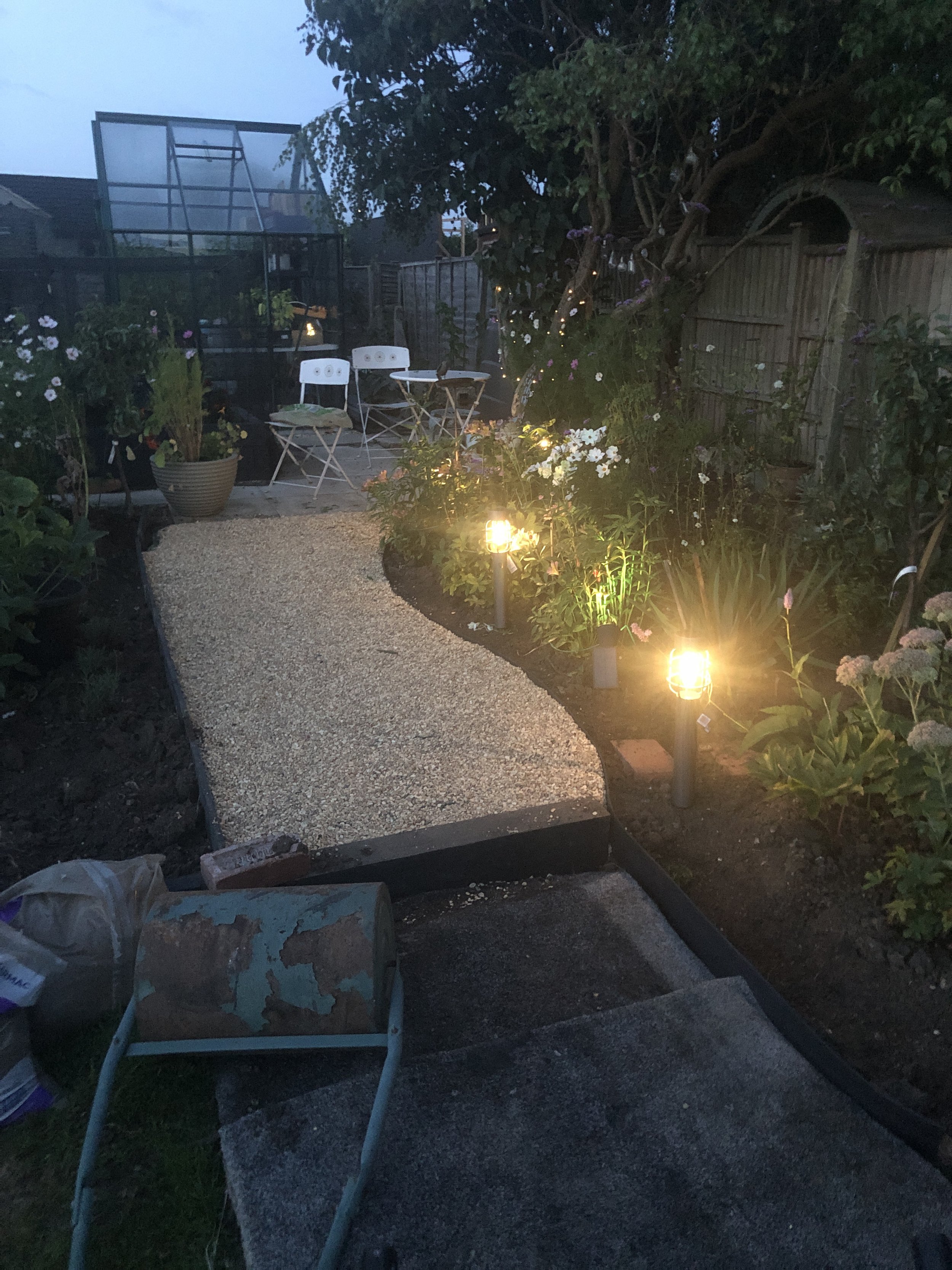 A backyard garden during dusk with a newly laid gravel pathway, potted plants, flower beds, a small white table with chairs, a tree wrapped with fairy lights, and outdoor lighting illuminating the area.