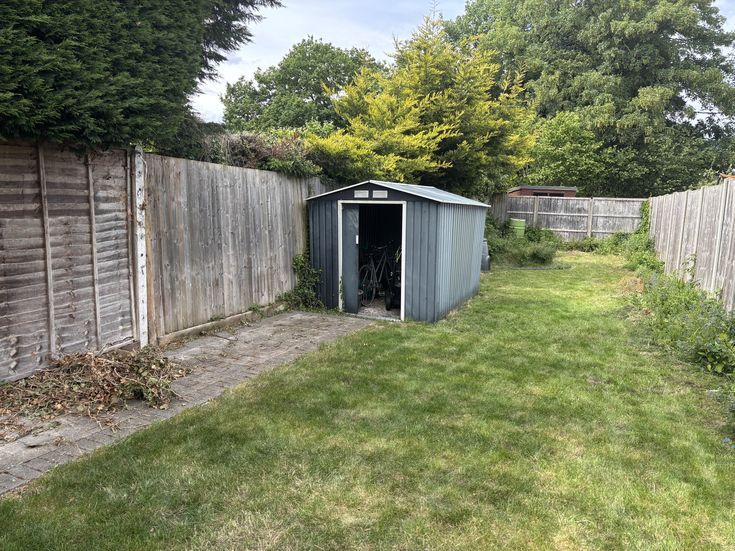 Backyard with a gray shed, wooden fence, green grass, trees, and a partially cloudy sky.