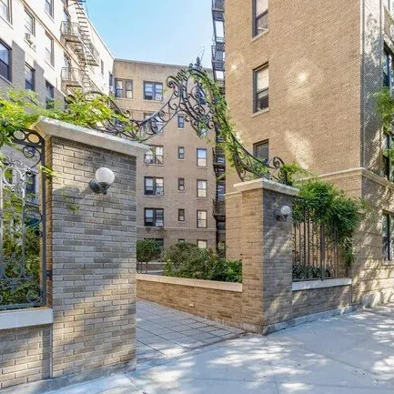Brick entrance gate with decorative black metalwork arch and street lamps, around a residential building complex with trees and sidewalk