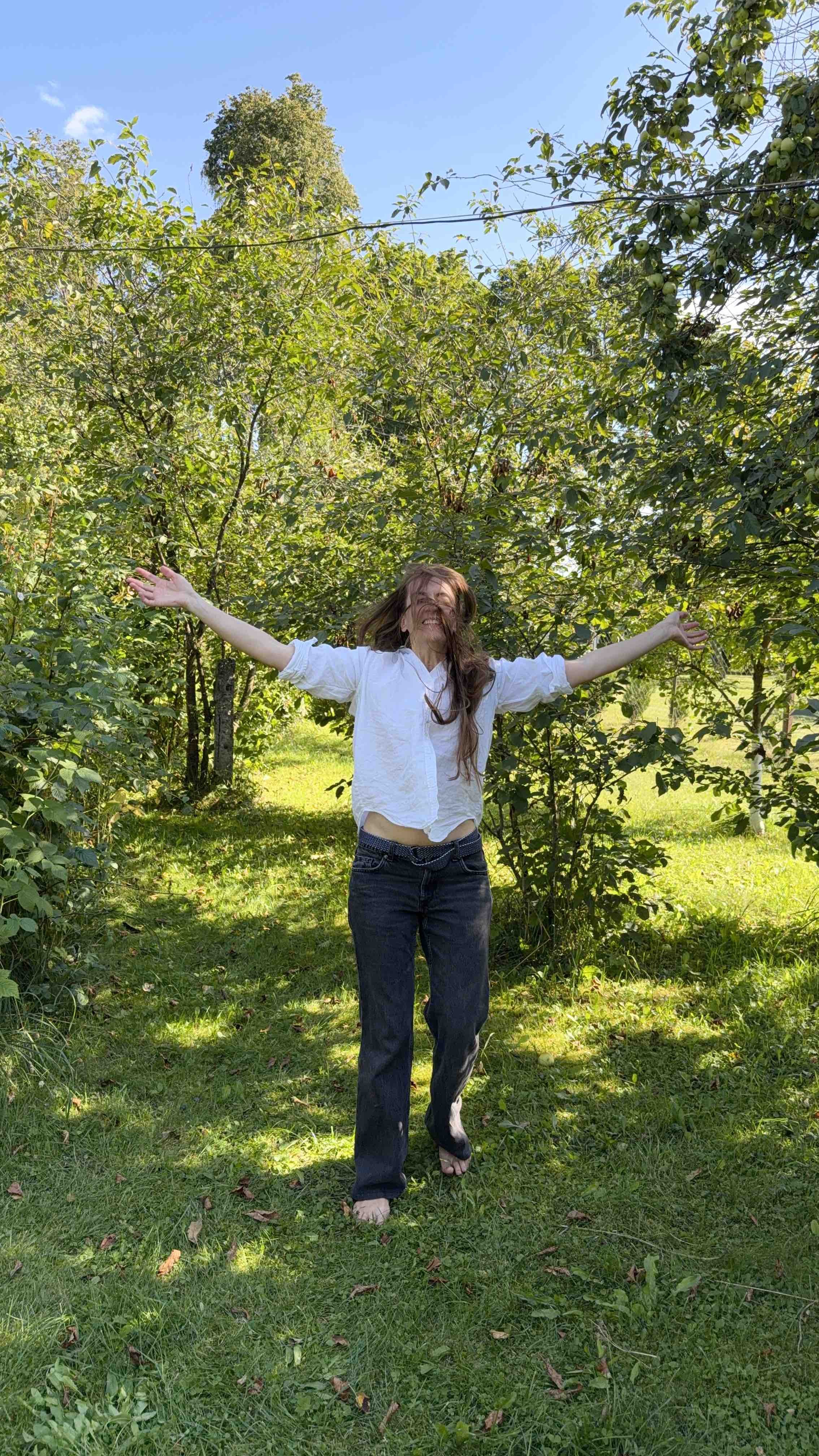 A woman with long hair wearing a white shirt and dark jeans, barefoot, standing in a grassy area with trees and green foliage, arms outstretched, smiling with her hair blowing in the wind on a sunny day.