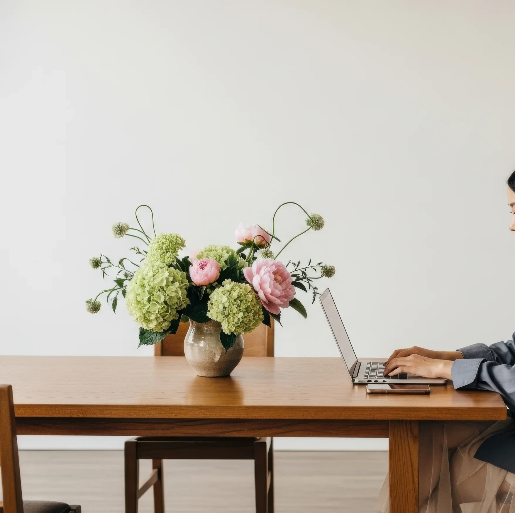 A person in a gray blazer working on a laptop at a wooden table with a flower arrangement in a beige vase on the left side.