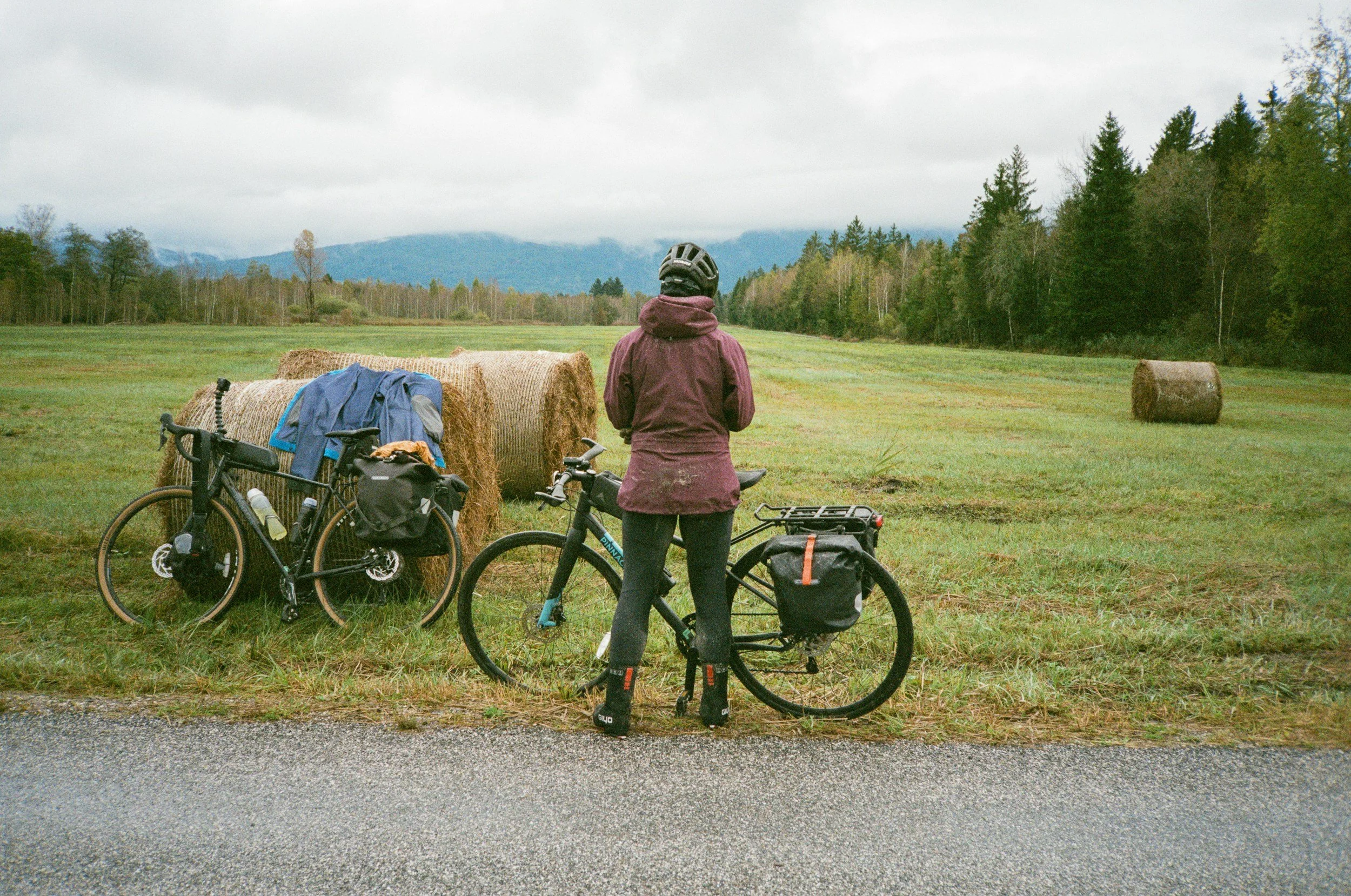 A person with a helmet and purple jacket standing next to two bicycles, facing a field with hay bales, trees, and mountains in the background during an overcast day.