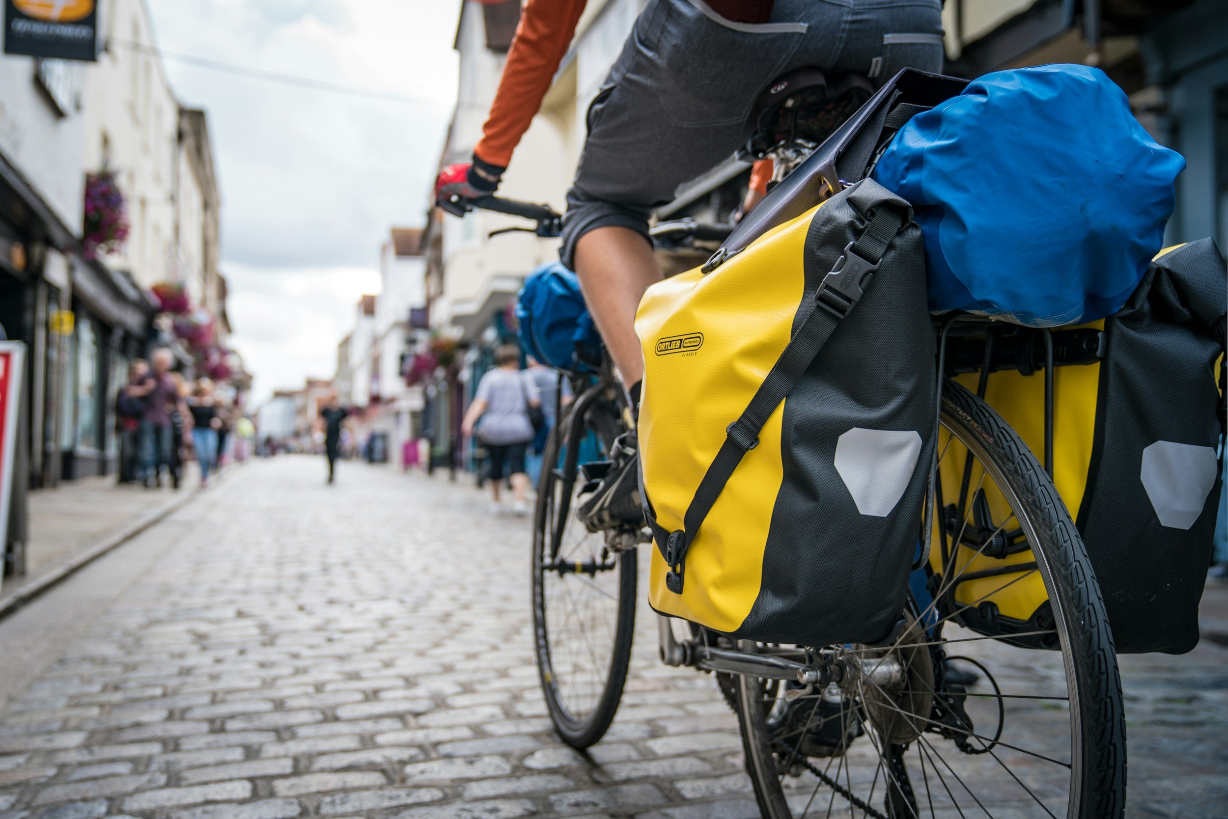 A bicycle with colorful panniers parked on a cobblestone street with shops and pedestrians in the background.