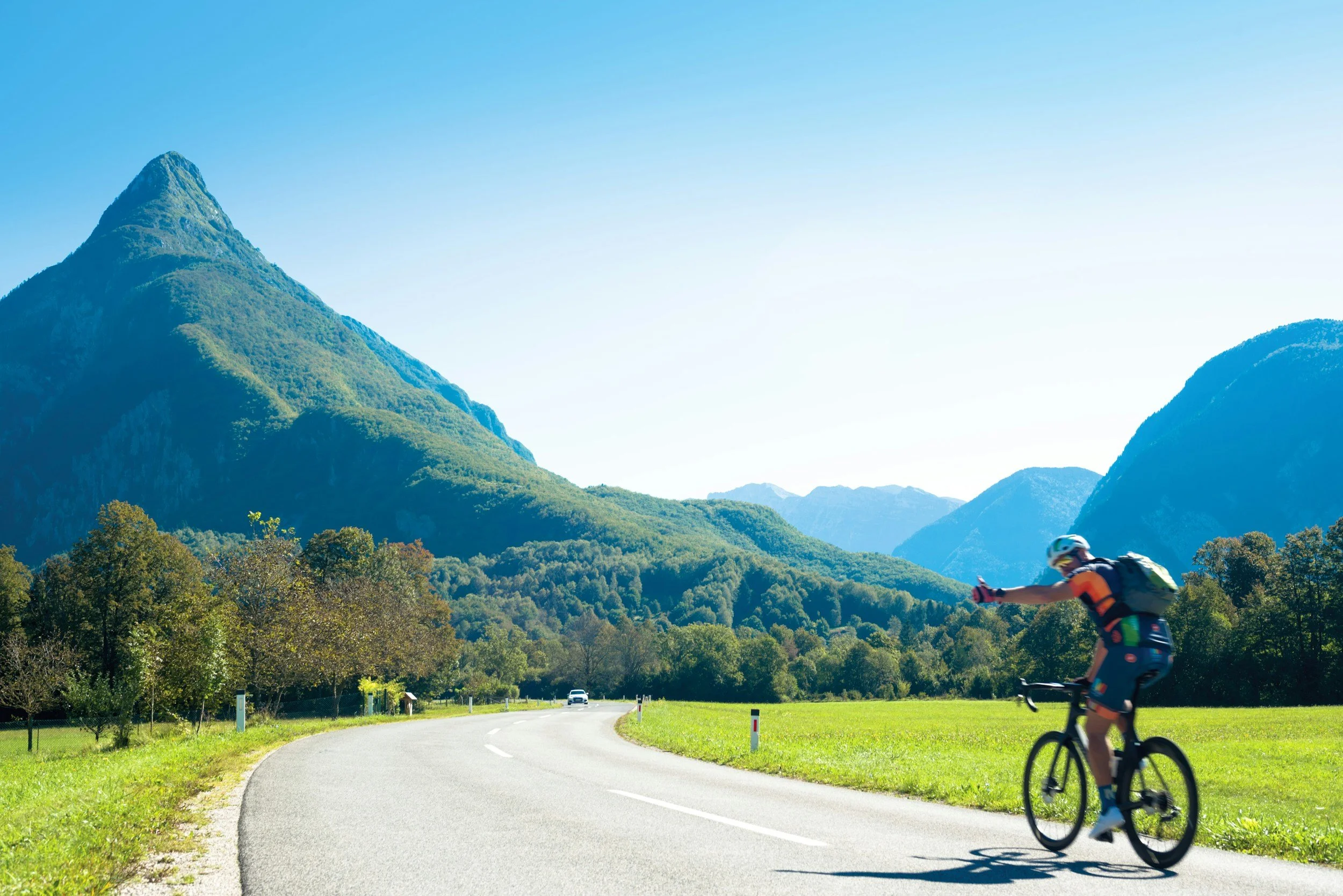Cyclist riding on a winding mountain road surrounded by green trees with mountains in the background under a clear blue sky.