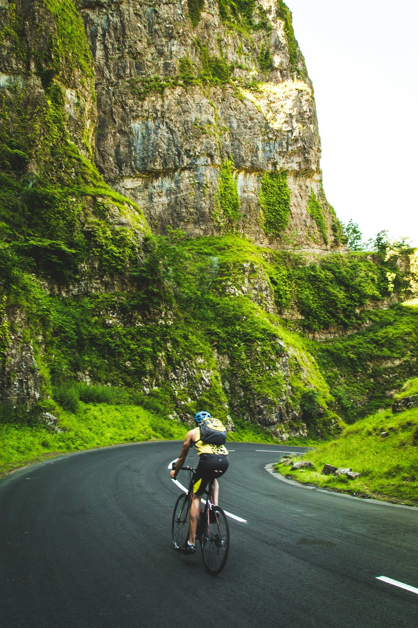 A cyclist wearing a blue helmet and yellow and black athletic clothing rides on a winding black asphalt mountain road surrounded by lush green vegetation and tall rocky cliffs.