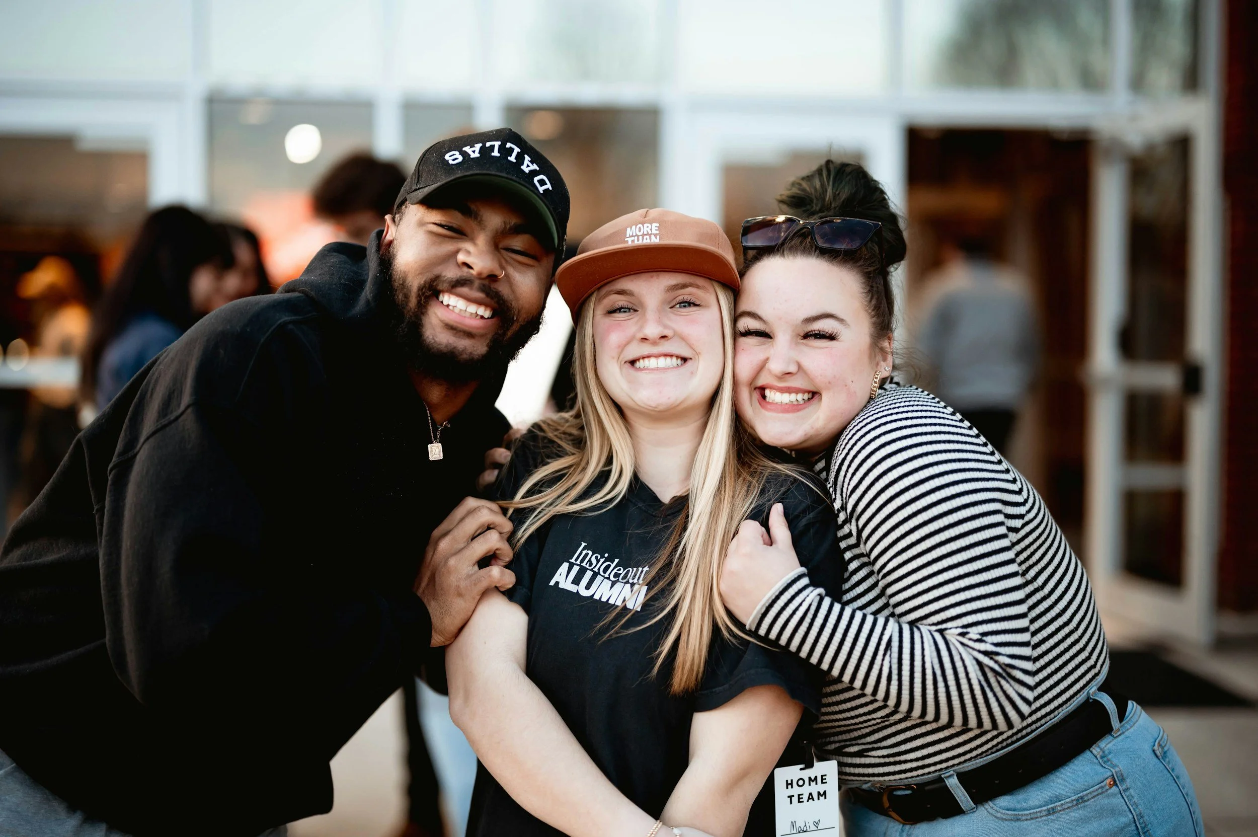 Three young people smiling and hugging each other indoors, with two women and one man, at a social gathering or event.