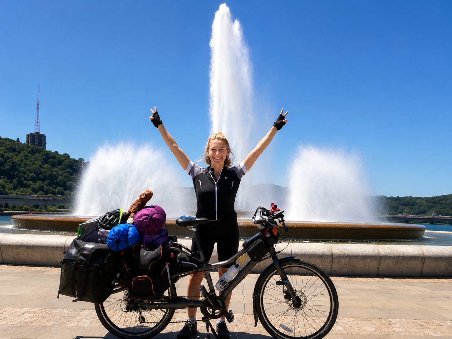 A woman smiling with arms raised doing a peace sign, standing in front of a fountain, with her loaded touring bicycle in front of her, outdoors on a bright sunny day.