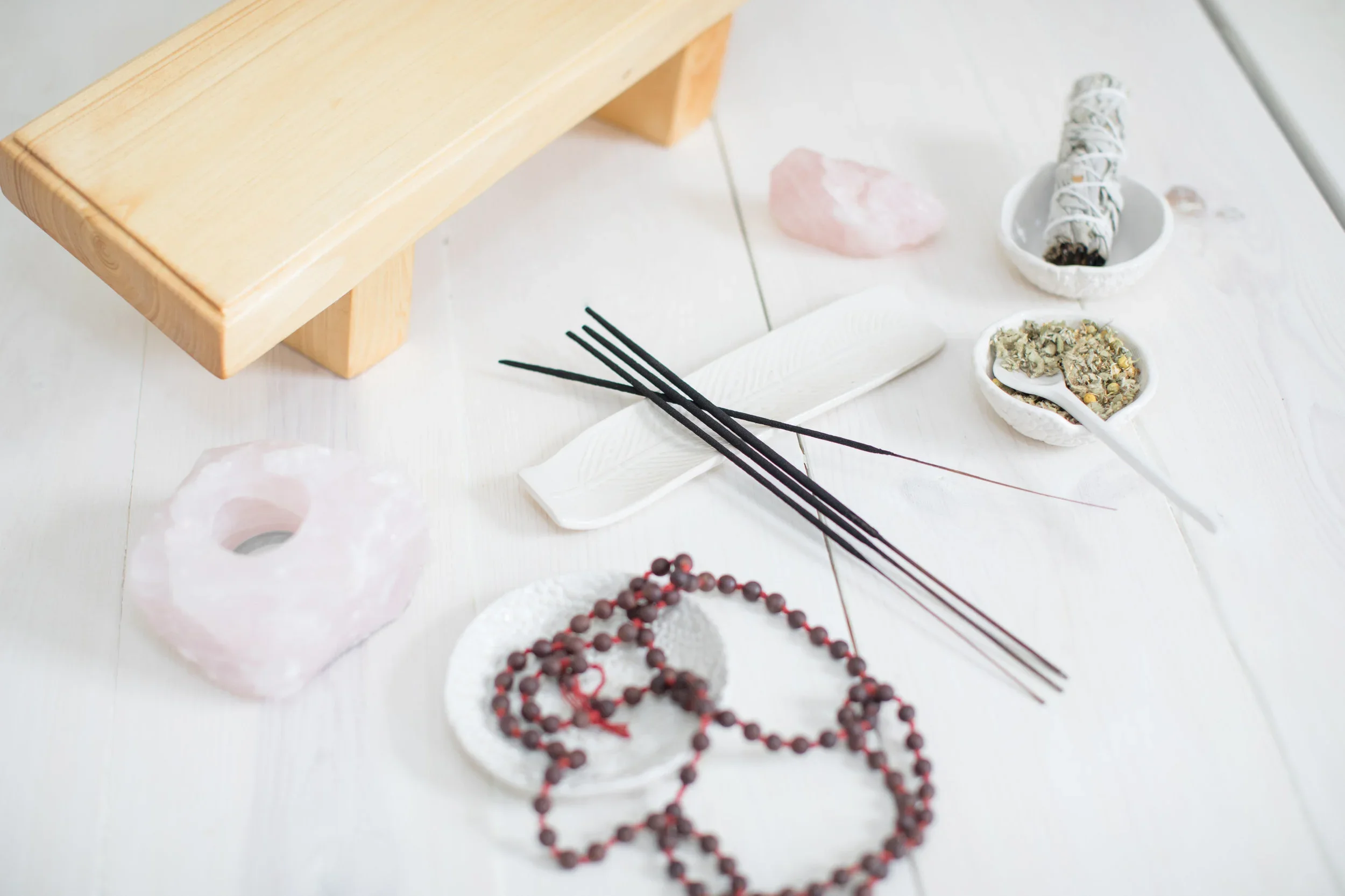 Decorative setting with a wooden stand, pink stone, white feather, black incense sticks, herbal mixture in small bowls, and a beaded necklace on a white surface.