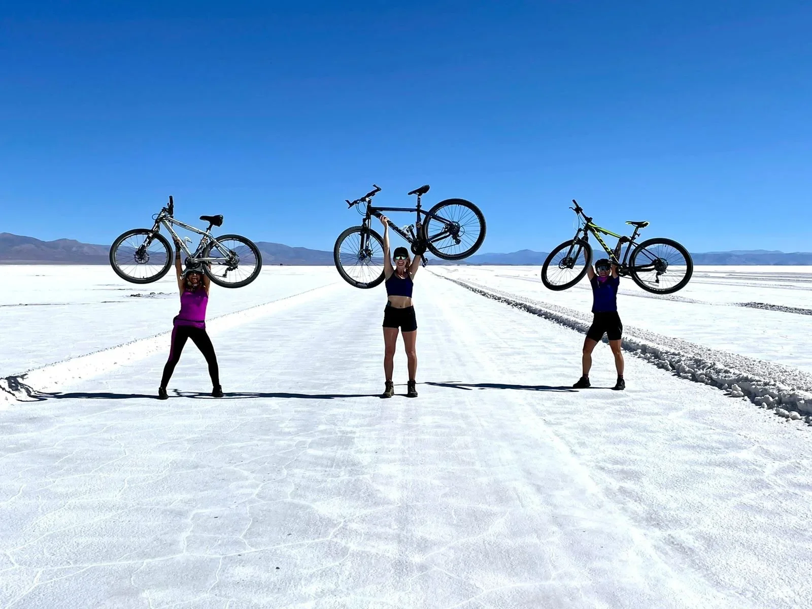Three women holding bicycles above their heads on a salt flat under a clear blue sky.