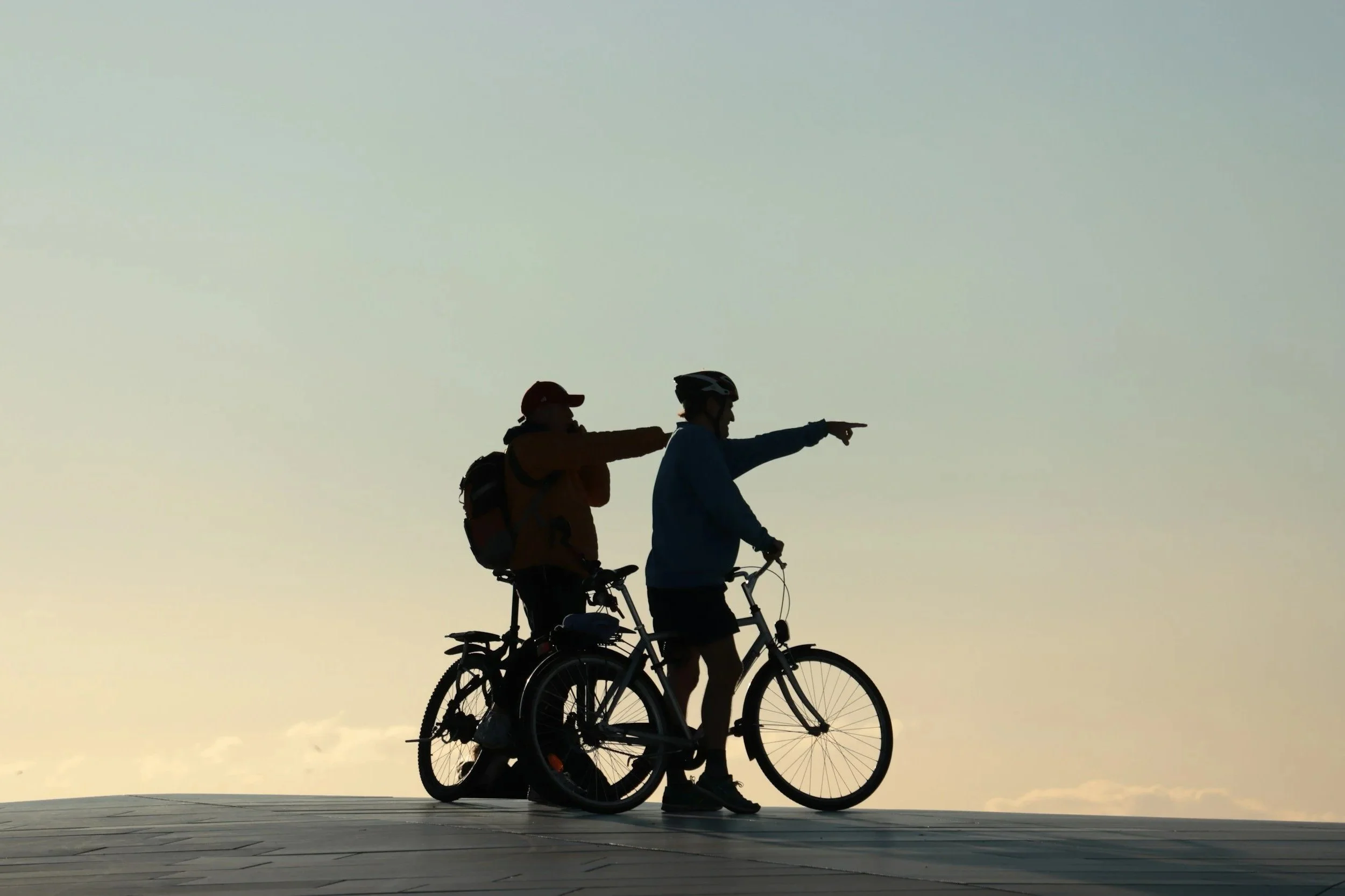 Two people riding bicycles at sunset, with one person pointing ahead, against a clear sky.