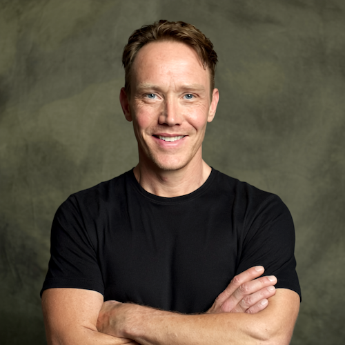 A smiling man with short brown hair, wearing a black t-shirt, standing with arms crossed against a greenish-gray background.