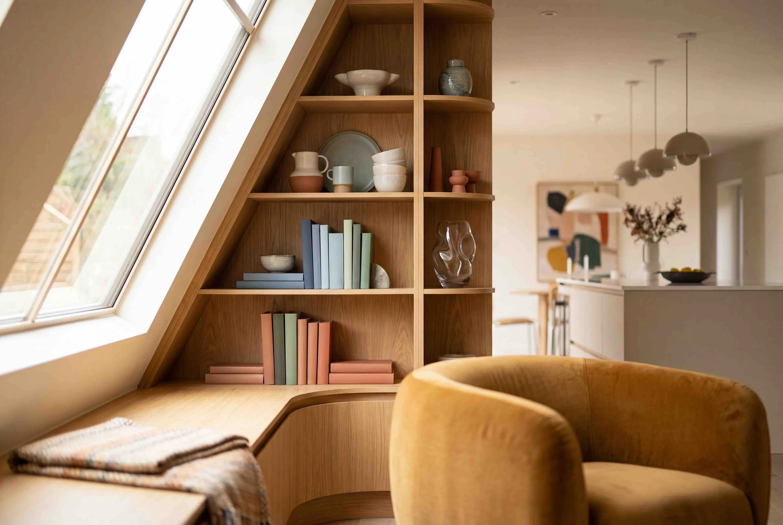 A wooden bookshelf near a large skylight window, with books, decorative items, and a glass vase, in an open modern kitchen and dining area.