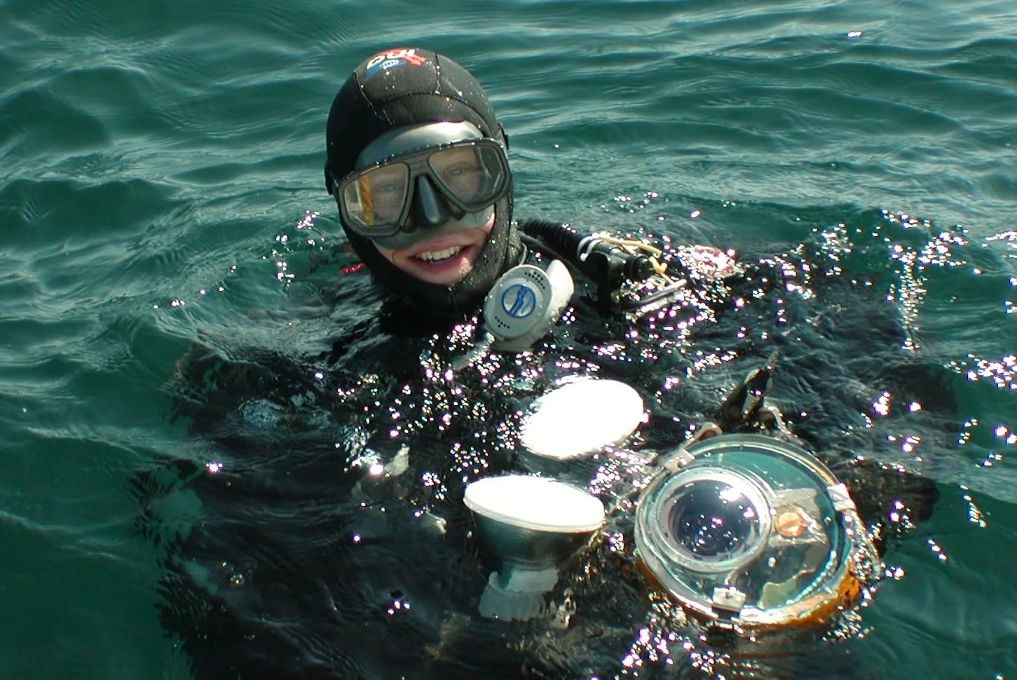 A person in scuba gear floating on the ocean surface, smiling and holding a piece of equipment underwater.