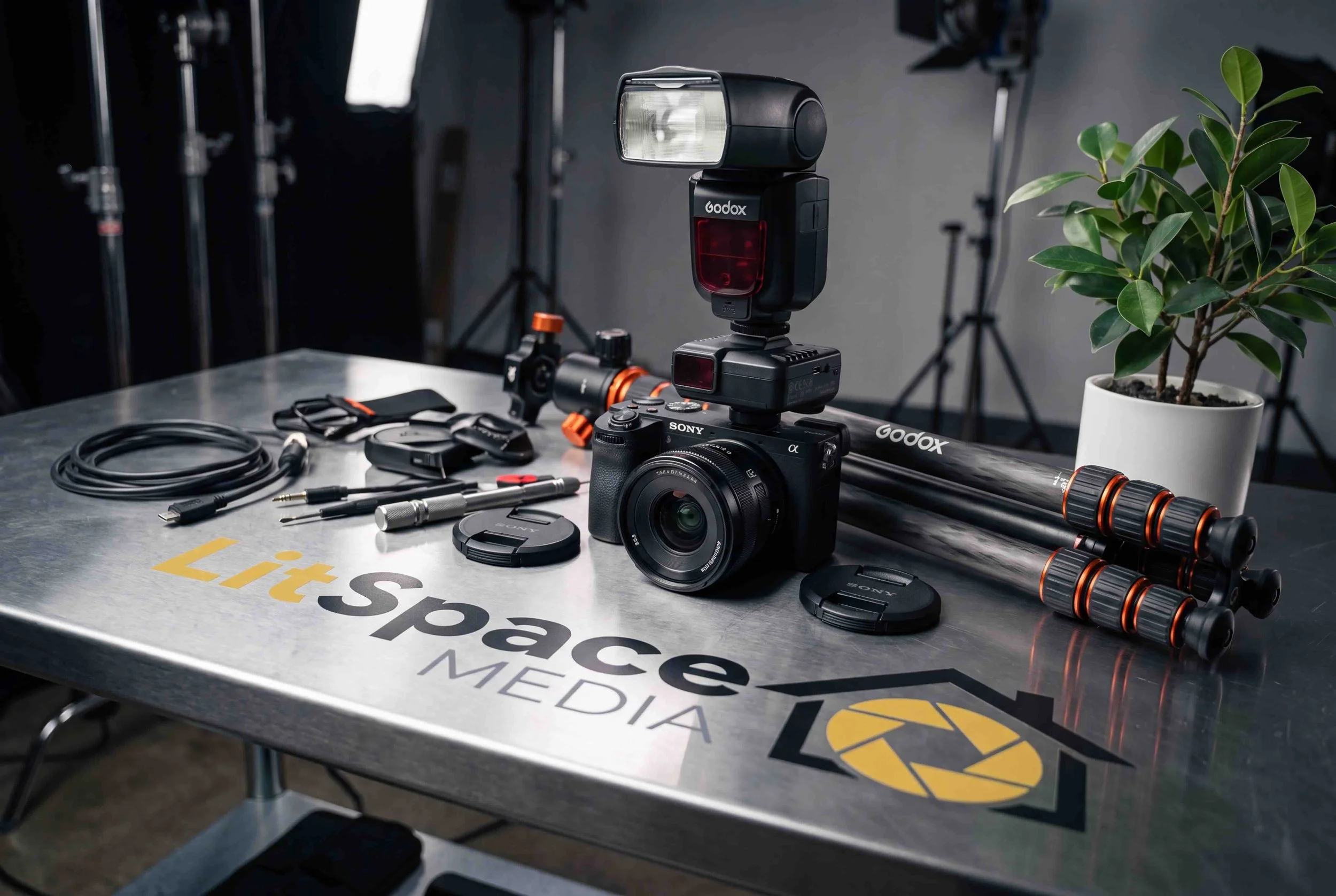 Upside-down camera with various camera accessories and lighting equipment on a metallic table with 'LiteSpace MEDIA' logo, a potted plant, studio lights, and tripods in the background.