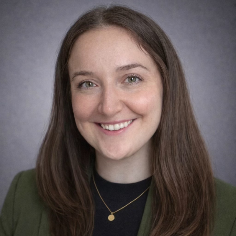 Close-up portrait of a young woman with long brown hair, smiling, wearing a black top, green blazer, and a gold necklace against a neutral background.