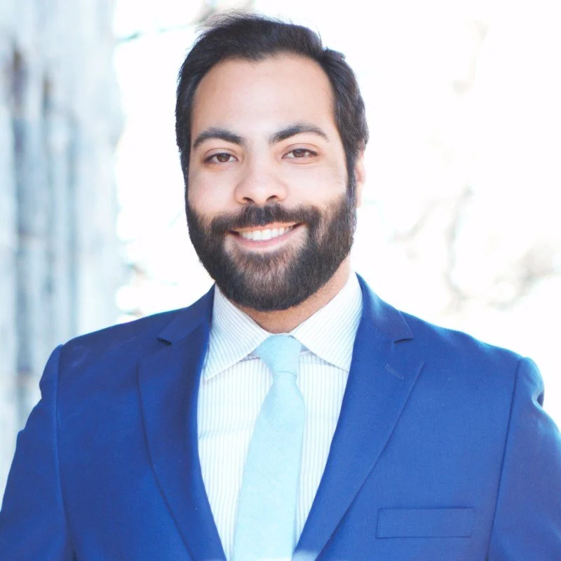 Professional man in a blue suit and light blue tie smiling outdoors.