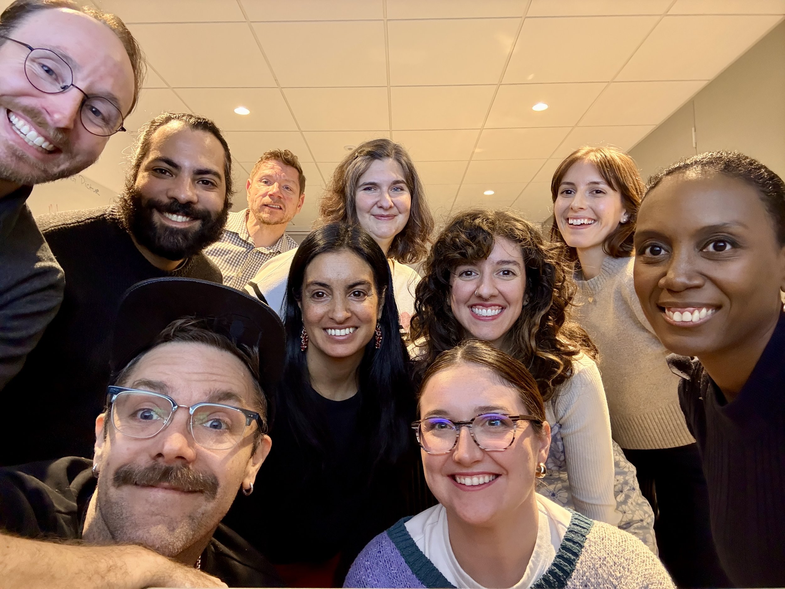 Group of diverse people smiling and posing for a selfie in an indoor setting, with a tiled ceiling and warm lighting.