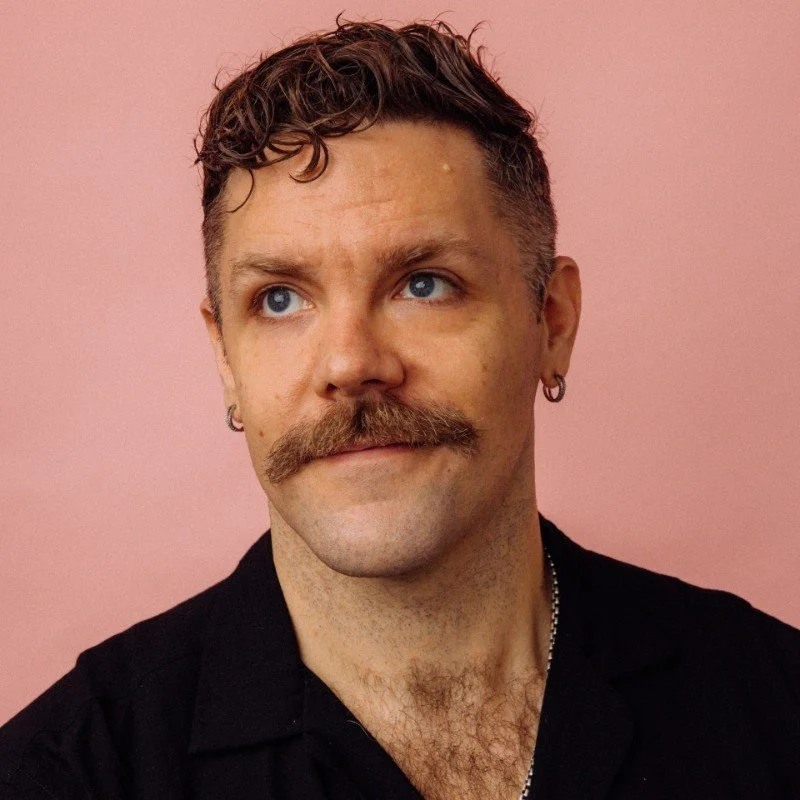 Close-up portrait of a man with brown curly hair, blue eyes, a thick mustache, and earrings, wearing a black shirt against a pink background.