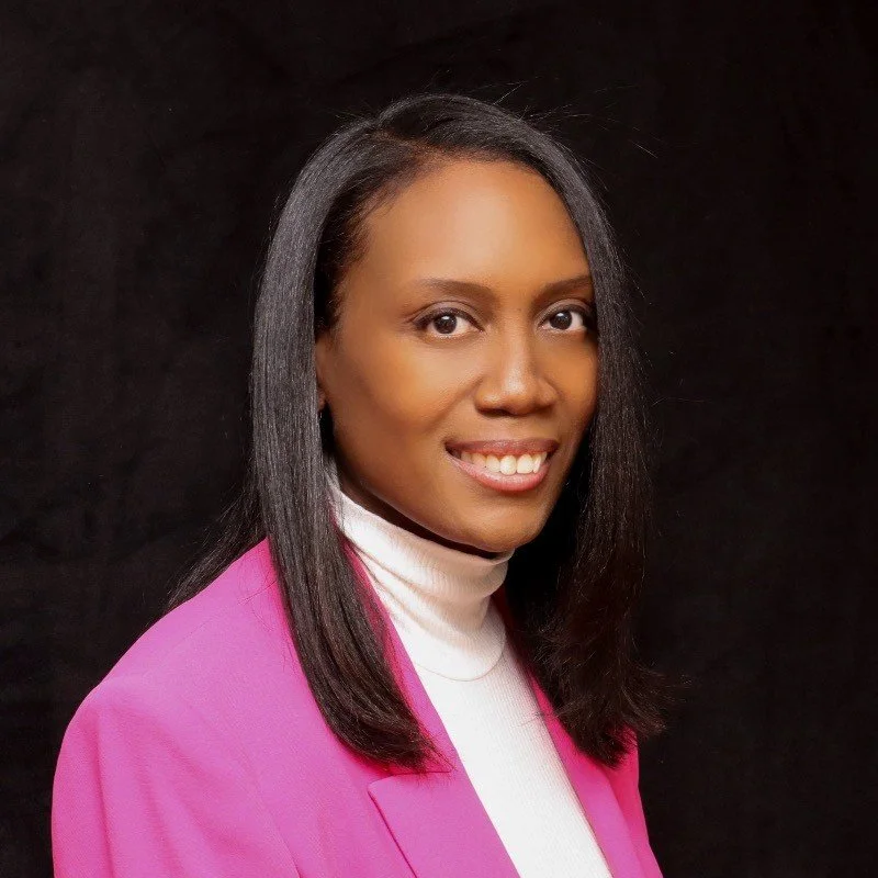 A young African American woman with straight black hair, wearing a white turtleneck sweater and a pink blazer, smiling against a dark background.