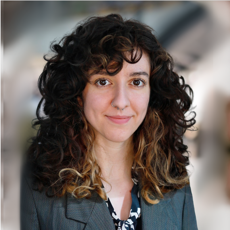 A woman with curly dark hair with lighter tips, wearing a gray blazer and a patterned blouse, smiling at the camera.