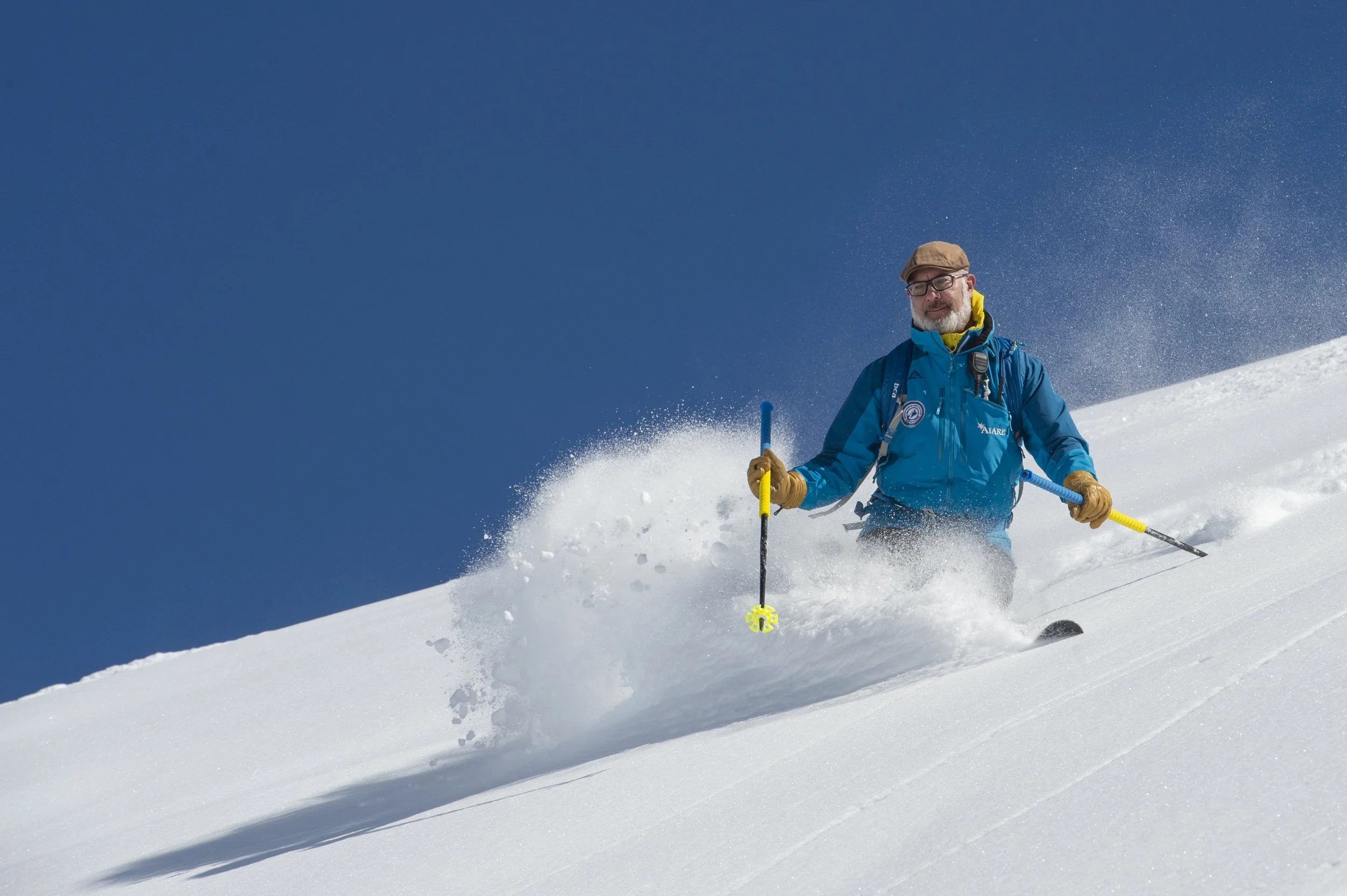 An older man with glasses, a brown cap, and a blue jacket skiing down a snowy slope under a clear blue sky.