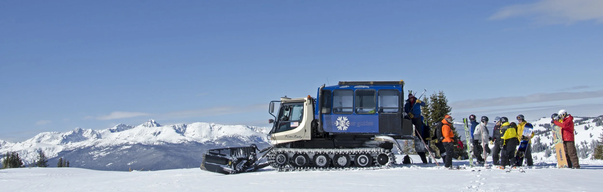 A snowcat vehicle with a group of skiers and snowboarders gathered on a snowy mountain with snow-covered peaks in the background.