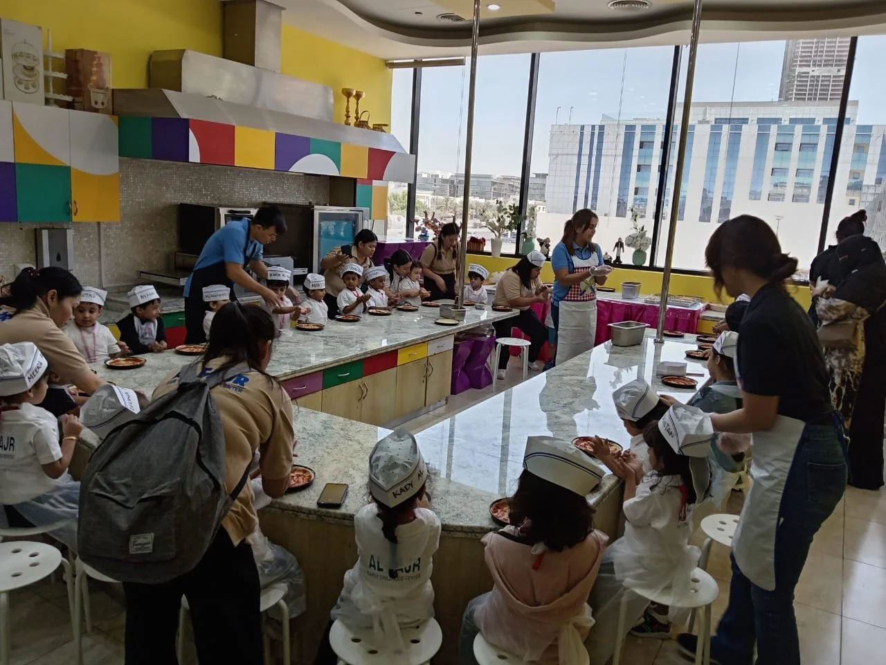 Children participating in a cooking class in a bright kitchen, wearing chef hats, with adults assisting, large windows in the background showing a cityscape.
