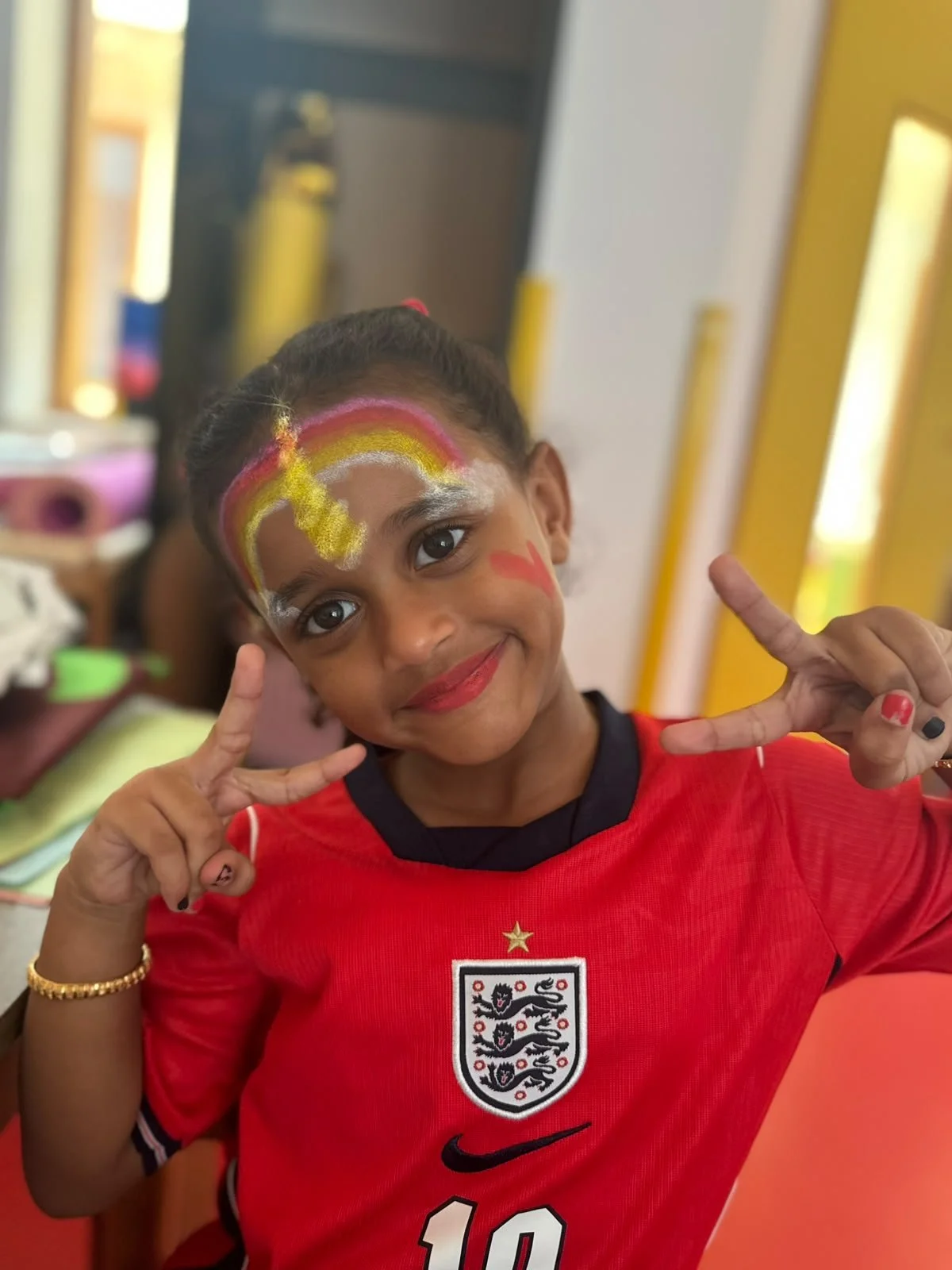 A young girl with face paint featuring a rainbow, white clouds, and a peace sign, smiling and making peace signs with her hands, wearing a red England football jersey.