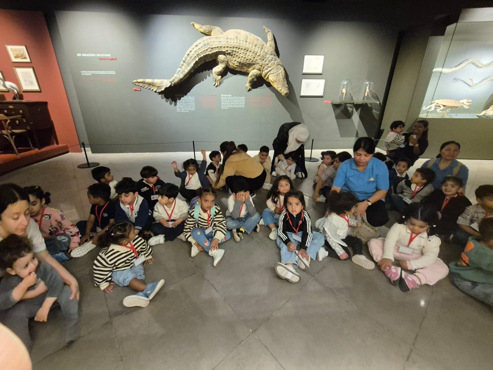 A group of children sitting on the floor in a museum, looking towards the camera, with a large crocodile sculpture mounted on the wall behind them.
