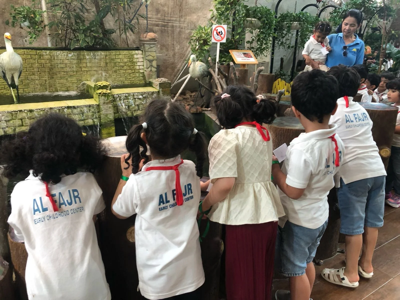 Group of children wearing white shirts with 'ALFAJR EARLY CHILDHOOD CENTER' on the back, gathered around an exhibit with waterfowl and water features, while a woman holds a child nearby, at an indoor zoo or nature exhibit.