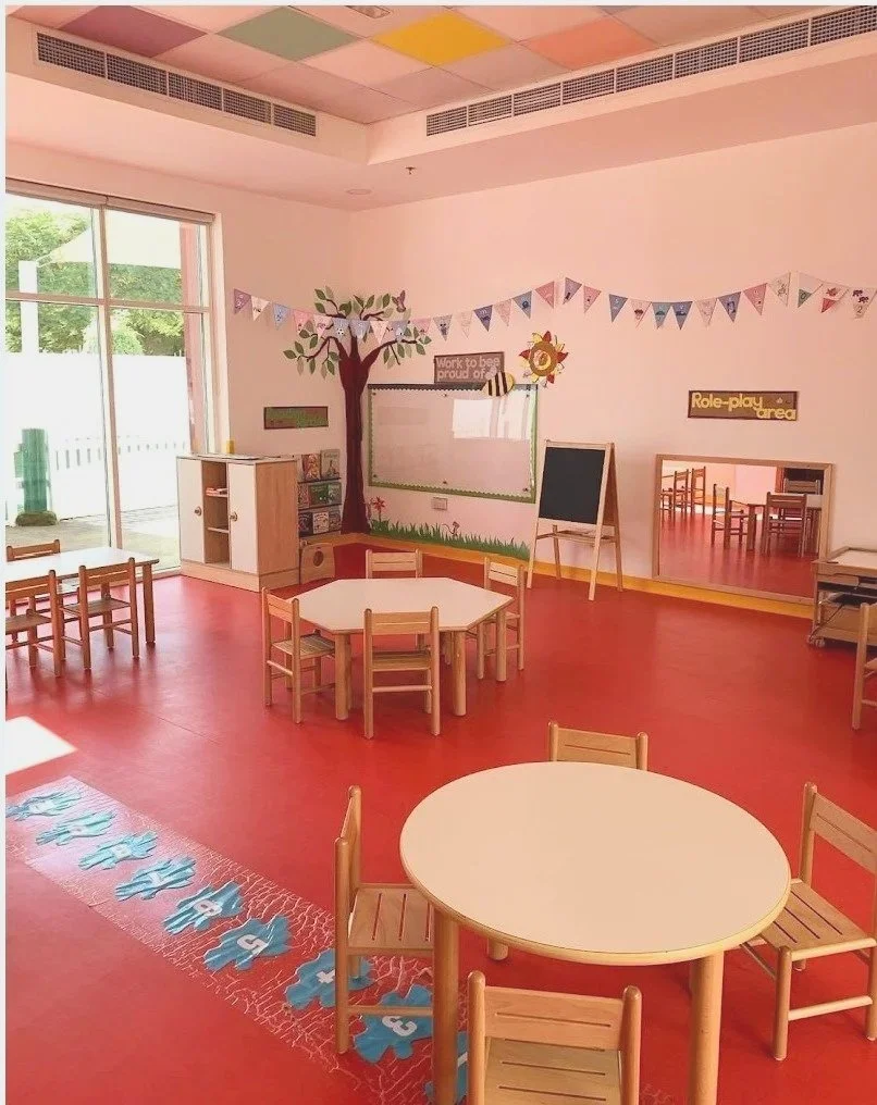 Colorful classroom with round tables, small chairs, a blackboard, and educational decorations, including a tree mural and bunting banners, with large window letting in natural light.