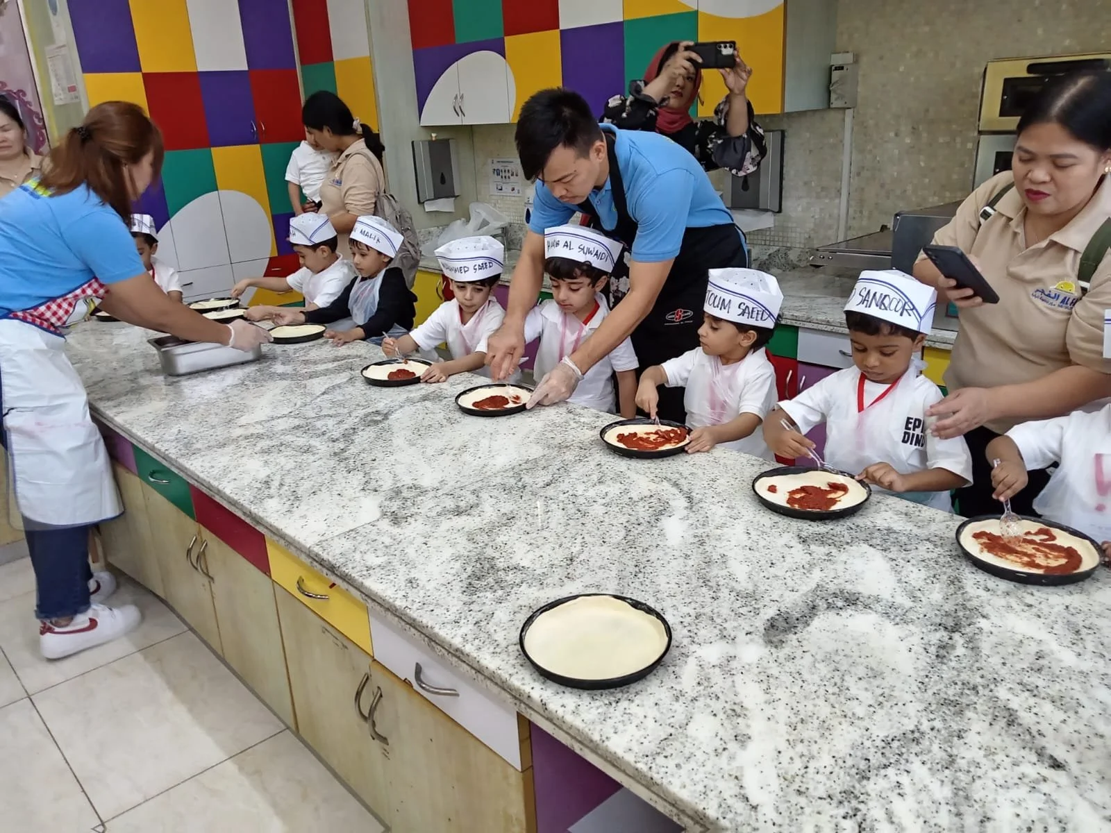 Children and adults preparing pizzas in a kitchen classroom. The children are wearing chef hats with their names, and the adults are assisting them with the pizza making process.