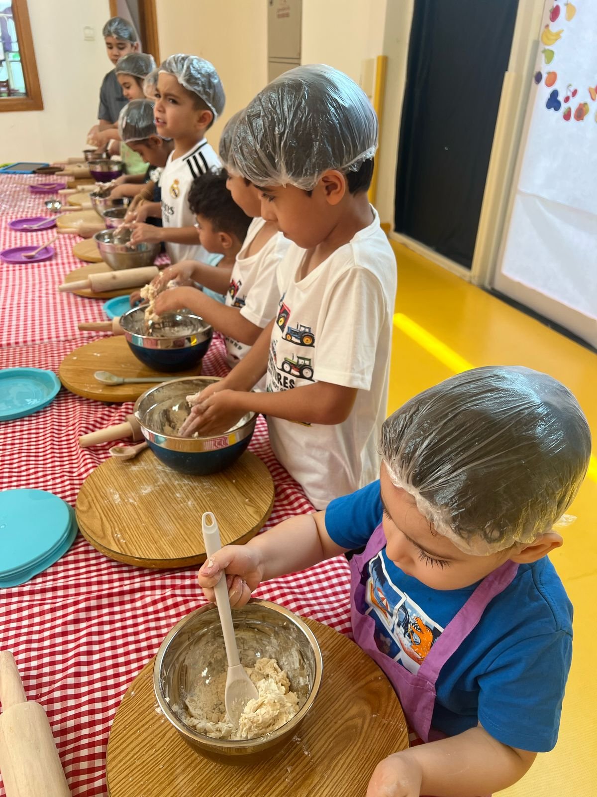 Children wearing hairnets and aprons participating in a baking activity at a long table with mixing bowls and rolling pins.