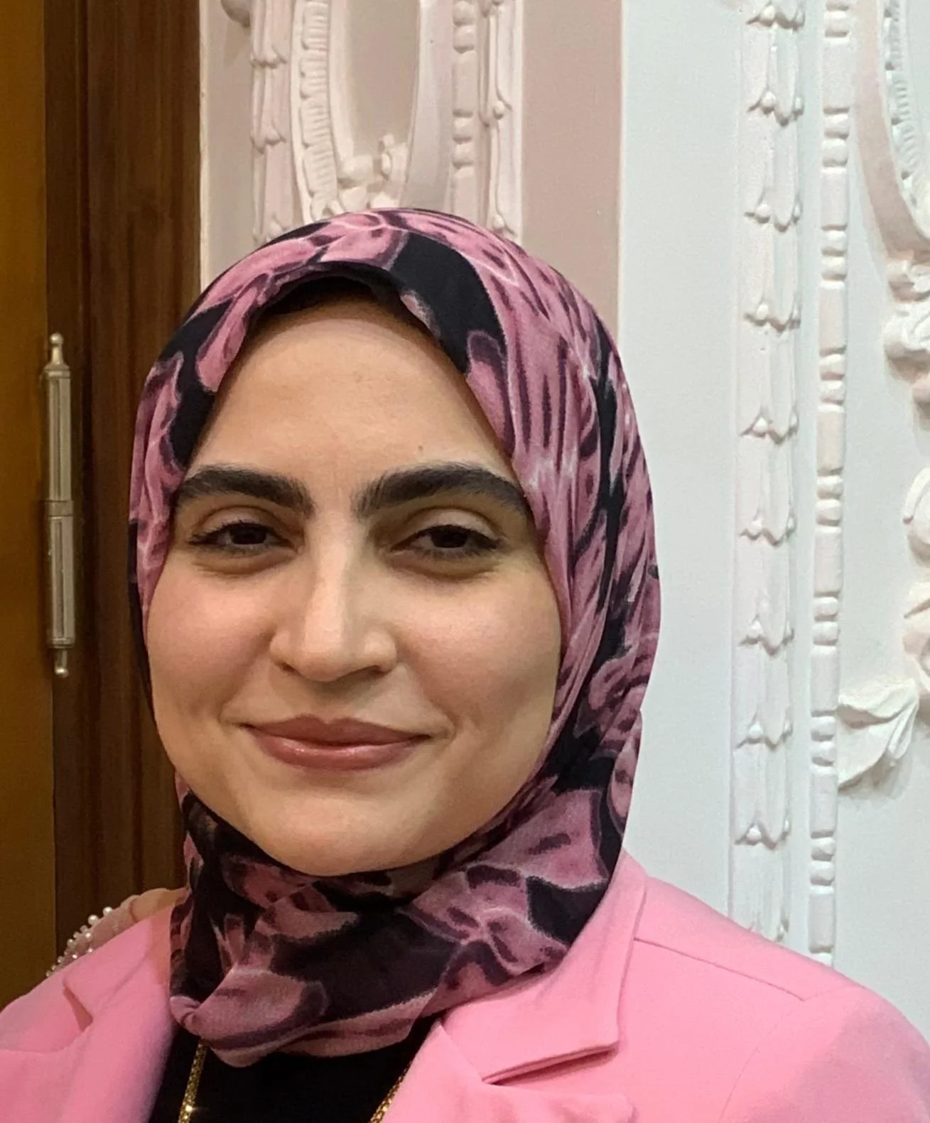 A woman with light skin wearing a pink blazer, a black shirt, a gold necklace, and a pink and black patterned headscarf. She is smiling slightly. The background features a white ornate decorative wall panel and a wooden door.