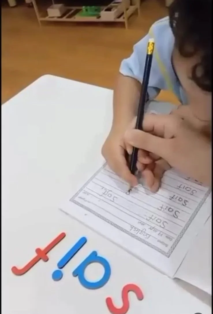 A young child writing a list titled 'SOFT' on a worksheet at a desk, with colorful letters spelling 'J!O'S' on the table in front.