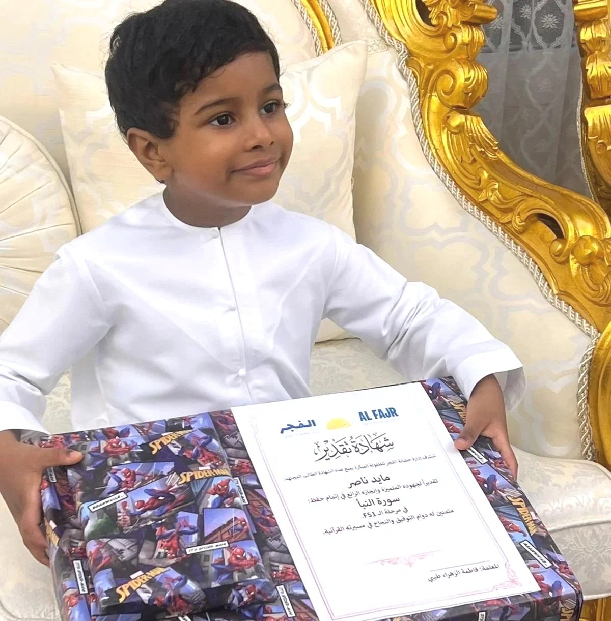 A young boy in traditional white attire sitting on a cream-colored sofa holding a colorful Spiderman-themed gift box with a certificate on top, in an ornate gold-embellished room.