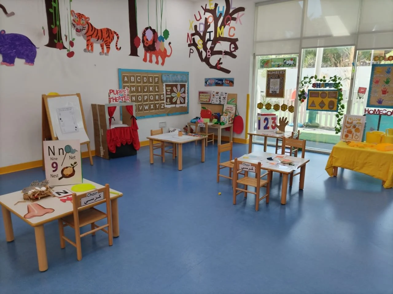 An early childhood classroom with small tables and chairs, educational wall decorations including animals and alphabet displays, and learning materials for children.