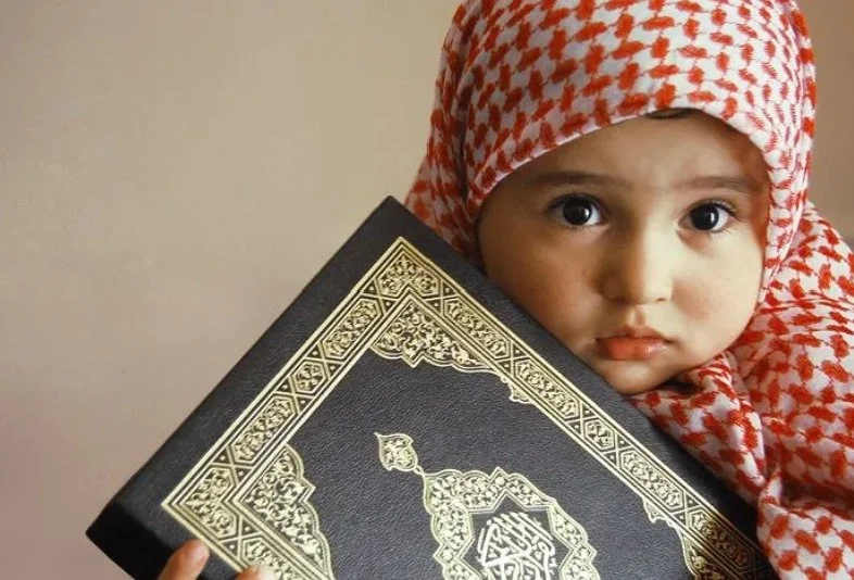 A young child with large eyes wearing a red and white checkered headscarf holding a black book with intricate gold Islamic geometric patterns.