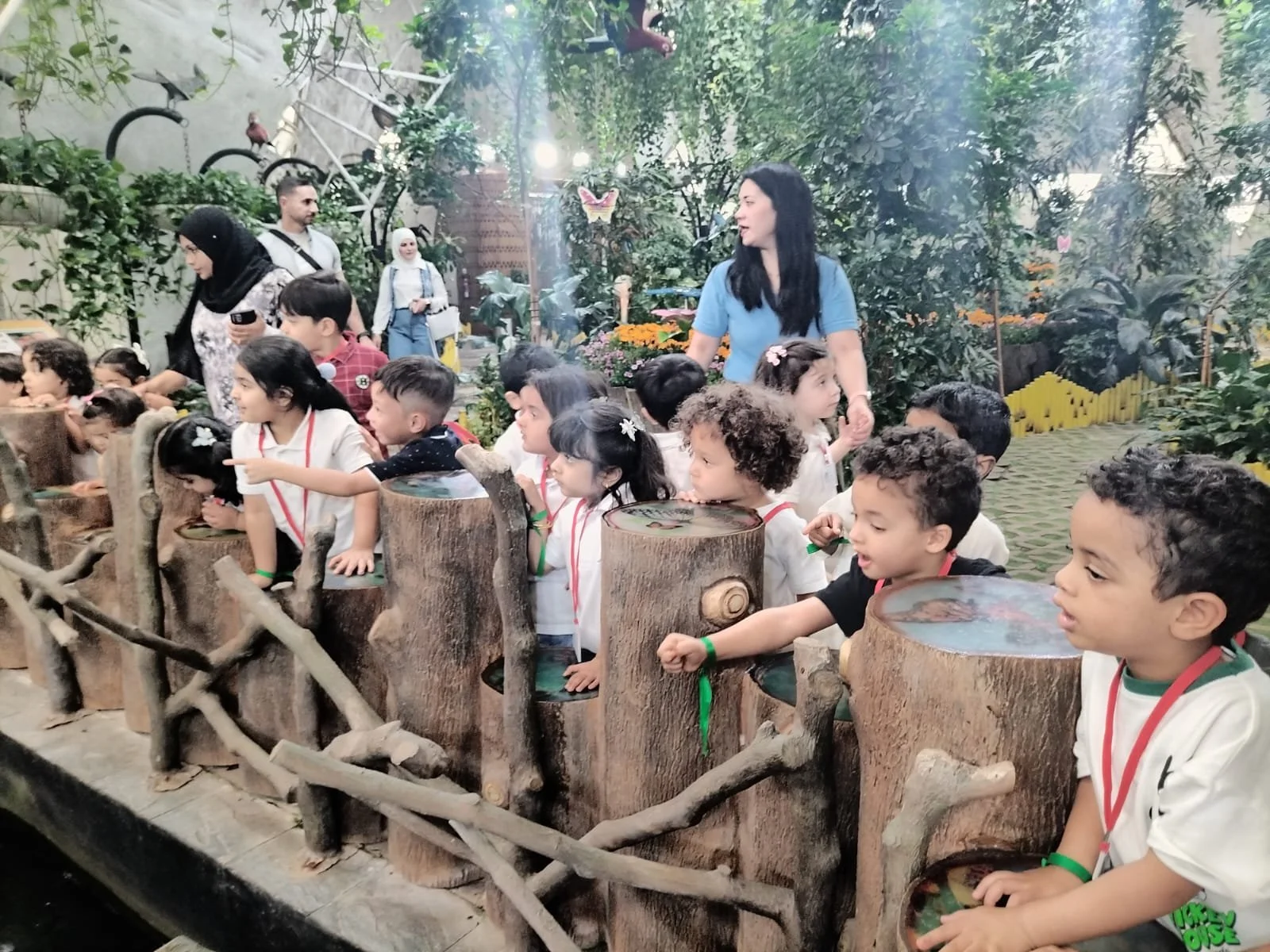 Children and adults visiting a lush, indoor botanical garden with plants, trees, and butterflies, with children leaning over logs as part of an educational activity.