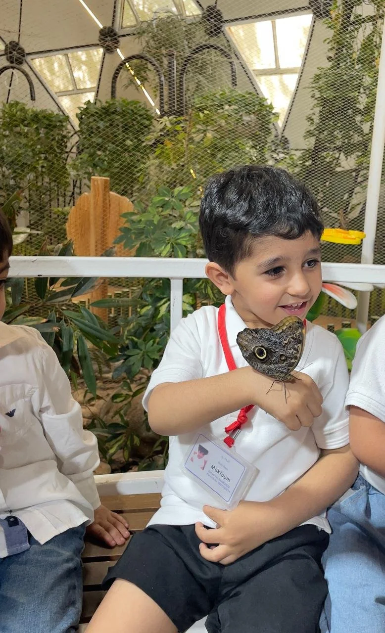 Young boy holding a butterfly on his hand at an indoor butterfly exhibit, smiling and wearing a name tag.