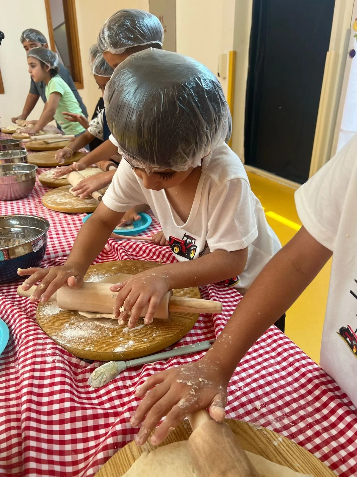 Children wearing hair coverings and aprons, rolling out dough with rolling pins on a table with a red checkered tablecloth in a classroom or kitchen setting.