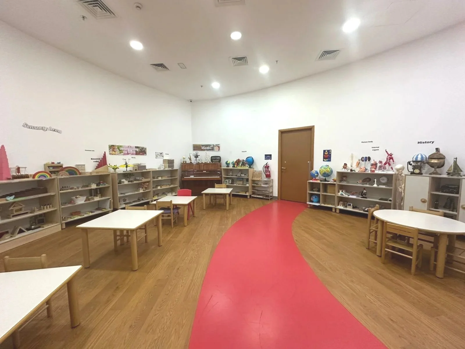 Children's classroom with wooden tables and shelves filled with educational toys and objects, a red carpet in the center, and white walls decorated with labels such as 'Sensory Area,' 'Music,' 'History,' and various educational displays.