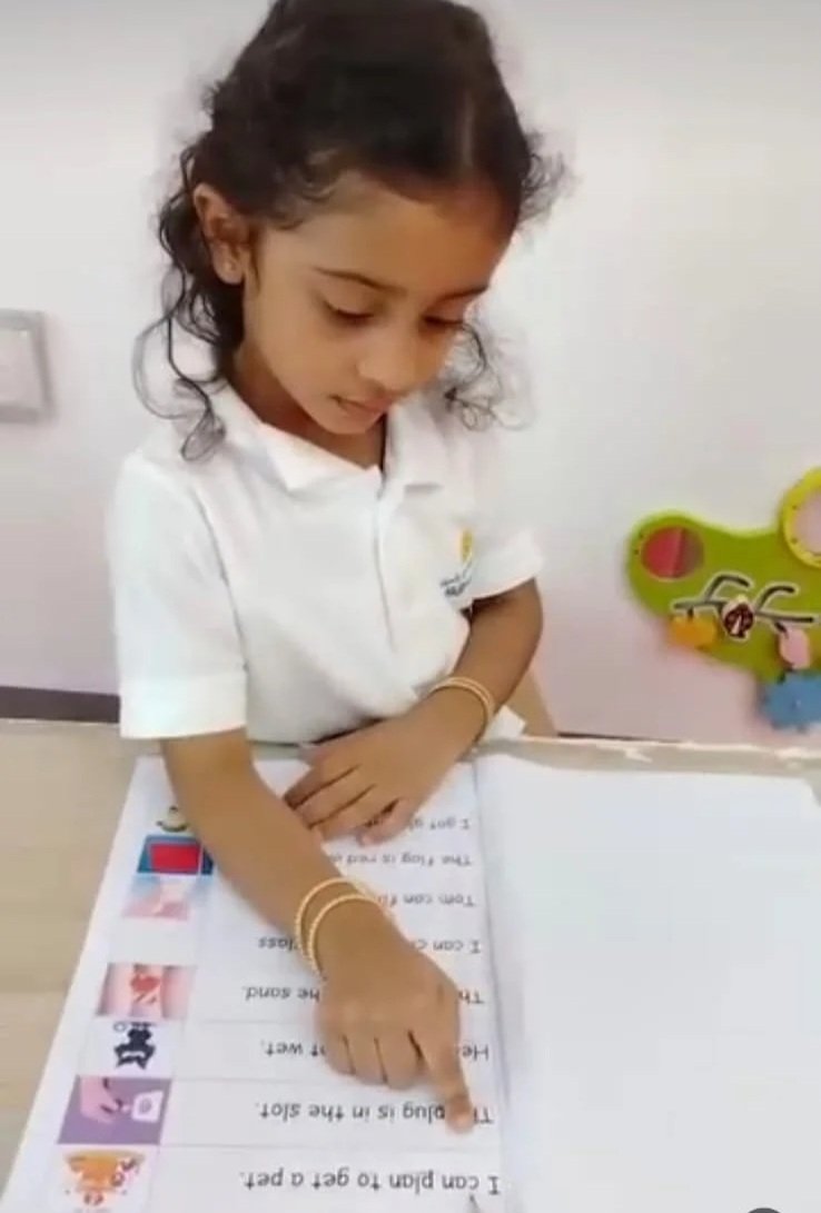 A young girl dressed in a white shirt with curly hair, sitting at a table pointing at a worksheet or paper with images and text.