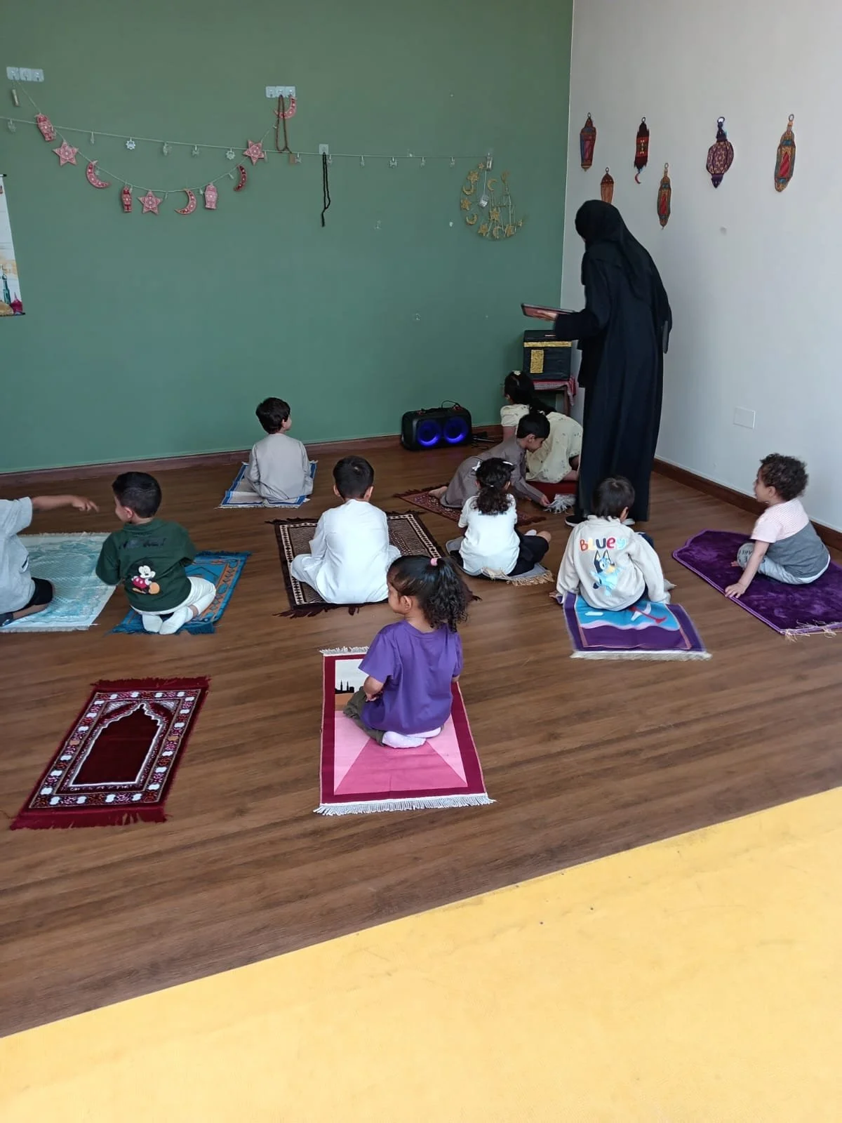Children sitting on prayer rugs in a classroom, listening to a woman in a black abaya.