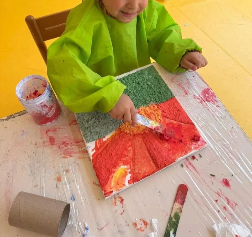 A young child wearing a bright green smock sitting at a painting table, creating a colorful artwork with textured green, orange, and red paints on a square canvas, using a palette knife.